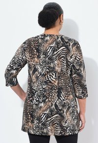 Frau mit dunklem Haar, das zu einem Dutt frisiert ist, trägt eine langärmlige Tunika mit Tierprint in Schwarz, Weiß und Braun, steht vor einem weißen Hintergrund.