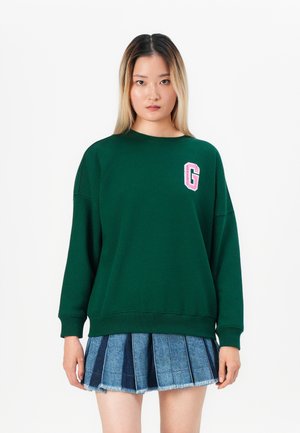 Green sweatshirt with a crew neck, dropped shoulders, and a pink 'G' logo. Paired with a pleated denim skirt featuring frayed edges.