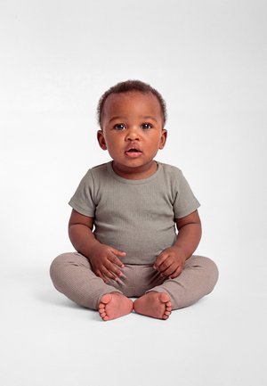 Zīdainis ar īsiem viļņotiem matiem, ģērbies pelēkā ribotā īssleju krekliņā un biksēs, sēžot ar krustu kājās uz balta fona.