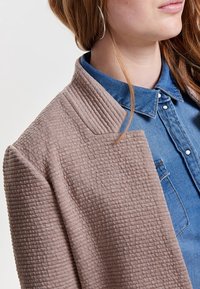 Vaalea beige tekstuurilla varustettu takki, jossa on hienovarainen tikattu kuvio, muotonsa mukainen kaulus ja avonainen etuosa, joka on puettu sinisen farkkutakin päälle.