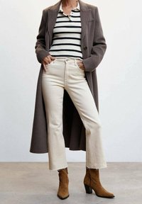 Largo abrigo marrón sobre una blusa de rayas en blanco y negro, acompañado de jeans acampanados color crema y botas de tobillo marrones con tacón.