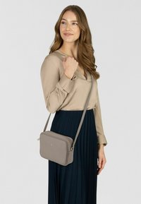 Bolso bandolera beige con textura de cuero suave, forma rectangular, cierre de cremallera y logo estampado en el frente. Correa ajustable.