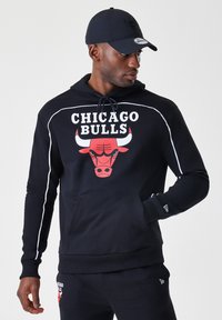 Crna dukserica s crvenim logom Chicago Bullsa, bijelim detaljima, podesivom kapuljačom sa vezicom i kengur džepom. Uparen s crnim hlačama.
