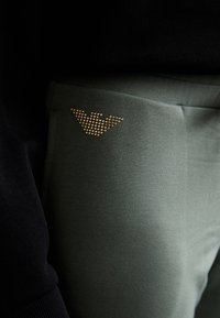 Gröna sweatpants har ett mjukt material, med en guldstrasslogotyp på framlappen och en svart topp som delvis syns ovanför.