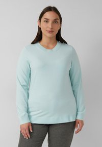 Vrouw met steil bruin haar, gekleed in een lichtblauwe trui met lange mouwen en donkere grijze broek, staande tegen een effen grijze achtergrond.