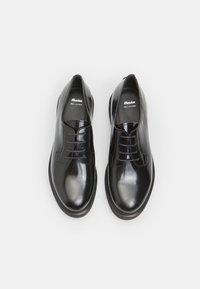 Chaussures en cuir noir avec une finition brillante, bout rond, à lacets et semelle plate en caoutchouc. Nom de la marque visible sur la semelle intérieure.