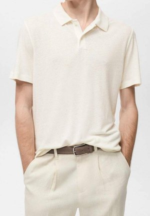 Homme portant un polo uni blanc cassé à manches courtes rentré dans un pantalon beige clair avec une ceinture marron, mains dans les poches.