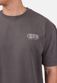 Šedé bavlněné tričko s kulatým výstřihem a krátkými rukávy, s bílým logem "CRYSTAL Paris" na levé straně hrudi.