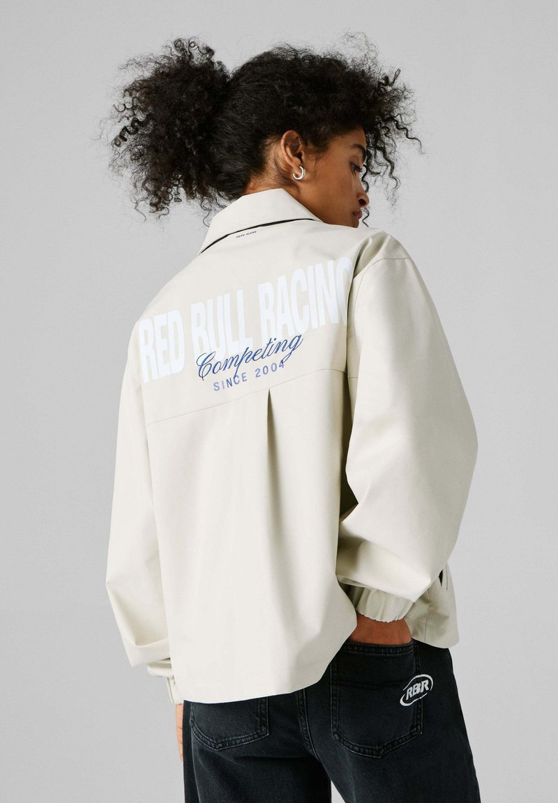Jonge persoon met krullend haar, gekleed in een licht jack met de tekst "RED BULL RACING Competing since 2004" op de achterkant en donkere broek.