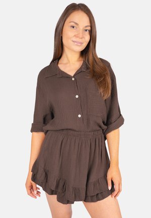 Jeune femme aux longs cheveux bruns portant une chemise boutonnée texturée marron foncé et un short assorti à volants, debout devant un fond uni.