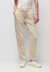 Beige Cordhose aus Kord mit Pattentaschen und Kordelzug in der Taille, getragen zu weißen Sneakern und einem in die Hose gesteckten weißen T-Shirt, stehend vor einer schlichten Wand.