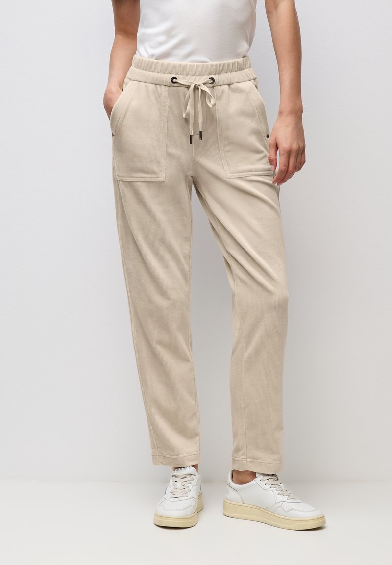 Beige Cordhose aus Kord mit Pattentaschen und Kordelzug in der Taille, getragen zu weißen Sneakern und einem in die Hose gesteckten weißen T-Shirt, stehend vor einer schlichten Wand.