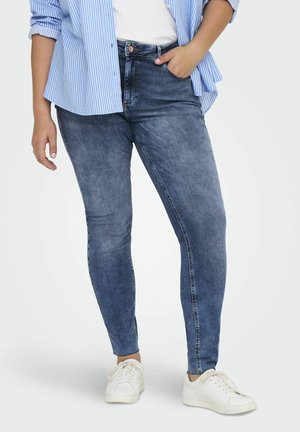 Skinny jeans in medium blue denim with a slight fade, featuring a high waist, button closure, and side pockets, paired with white sneakers.