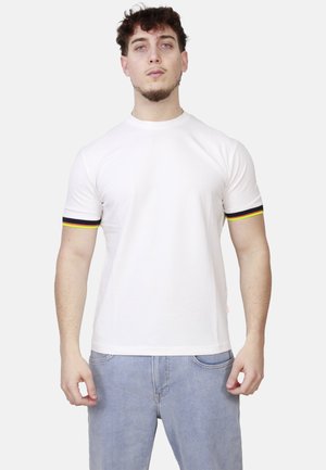Giovane uomo con capelli corti e ricci indossa una maglietta bianca con bordi neri, gialli e rossi sulle maniche e jeans azzurri su sfondo neutro.