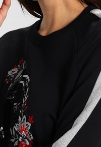 Sweatshirt noir avec un design floral brodé en rouge et gris, présentant des détails texturés et des accents de manches gris contrastants.