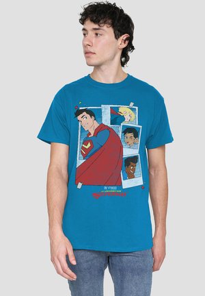 Young man with dark curly hair wearing a blue t-shirt featuring animated Superman and three other characters in comic-style frames.