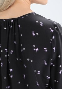 Černá blůza s malými fialovými květinovými vzory, s jemným nařasením kolem výstřihu a hladkým, lehkým materiálem.