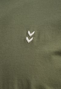 Chemise en tissu vert olive à la texture lisse, comportant deux lignes blanches brodées en forme de chevrons sur le côté gauche.