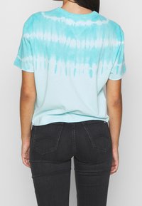 Mujer que lleva una camiseta de teñido anudado en azul claro y blanco y jeans ajustados negros, vista desde atrás contra un fondo sencillo.