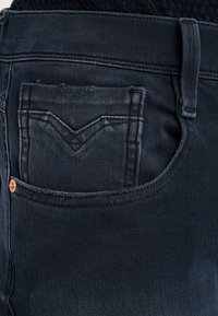 Mörka denimjeans med en bakficka som har en distinkt V-formad sömdesign. Kopparnitar nära midjan.