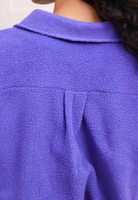 Fialová fleecová bunda s měkkou texturou, klasickým límcem a malým záhybem na zádech. Bez viditelných kovových doplňků nebo vzorů.