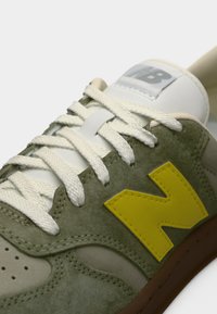 Gros plan sur une chaussure de sport en daim vert avec des lacets blancs, un logo "N" jaune sur le côté et une languette blanche portant une inscription partielle "NB".