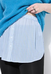 Maglione blu lavorato a maglia indossato sopra una camicia a righe bianche e azzurre con orlo arrotondato, dotata di dettagli con bottoni lungo la parte anteriore.