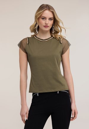 Junge Frau mit blondem Haar trägt ein olivgrünes Oberteil mit Perlenbesatz am Ausschnitt und transparenten Cap-Ärmeln, kombiniert mit schwarzen Hosen mit Reißverschlusstaschen.