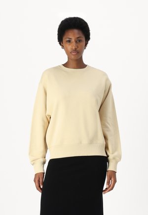 Beiger Pullover mit rundem Ausschnitt und gerippten Bündchen. Weiche Textur und lockere Passform, kombiniert mit einem schwarzen Rock.