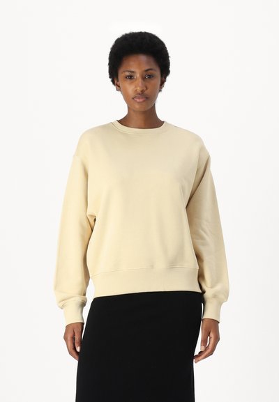 Sweatshirt bej cu guler rotund și mansete din material ribbed. Textură moale și croială lejeră, asortată cu o fustă neagră.