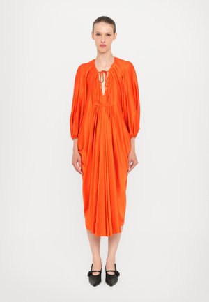 Heldere oranje jurk met een geplooide romp, diepe halslijn en pofmouwen. Lange, vloeiende silhoutte met een zachte textuur. Gedragen met zwarte schoenen.