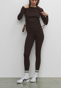 Bruin nauwsluitend shirt met lange mouwen en bijpassende legging van rekbare stof. Witte sneakers met zwarte accenten en gestreepte sokken. Springtouw in de hand.