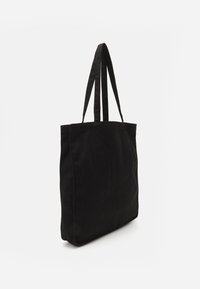 Sac fourre-tout noir en tissu, doté de deux longues poignées, d'une forme rectangulaire et d'un design simple sans accents ni motifs visibles.