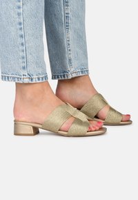 Goldene, gewebte Sandalen mit Absatz und Kreuzriemen-Design, niedrigem Blockabsatz und offener Zehe, kombiniert mit hellblauen, taillierten Jeans.