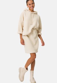 Felpa color crema, morbida e pelosa, con cappuccio regolabile e maniche arrotolate, abbinata a una gonna coordinata a ginocchio e stivali alla caviglia beige.