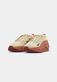 Beige athletic shoes with a textured surface, orange rubber sole, and black logo. Features lace-up design and pull tab at the heel.