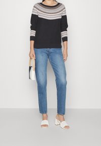 Svart stickad tröja med ett geometriskt mönster i vitt och beige vid halsringningen, i kombination med blå jeans och vita blockklackade mule-skor.