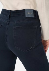 Jeans bleu foncé en tissu extensible, avec des poches arrière cousues, un patch logo à la taille et une texture lisse et élégante.