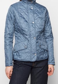 Jasnoniebieska pikowana kurtka z wysokim kołnierzem, zatrzaskami i dwoma przednimi kieszeniami z patkami. Gładka, błyszcząca faktura materiału.