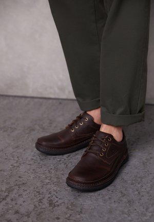 Braune Lederschuhe mit runden Zehen, Schnürdesign und dekorativer Naht. Kombiniert mit dunkelgrünen, hochgekrempelten Hosen.