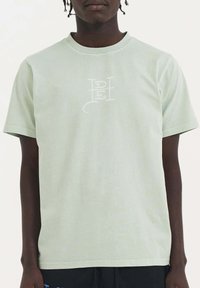 Camiseta de algodón verde claro con cuello redondo y mangas cortas. Presenta un logo blanco en el centro. Textura suave.