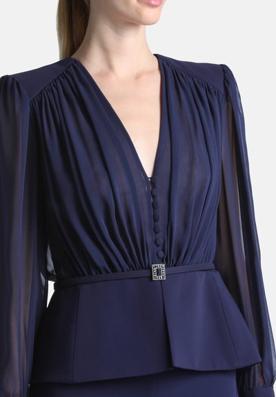 Femme portant une robe bleu marine avec des manches longues transparentes, un profond décolleté en V avec des boutons recouverts de tissu, et une boucle de ceinture décorative.