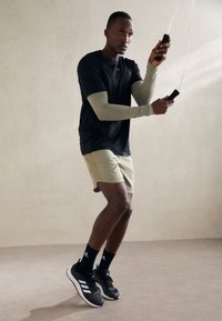 Black athletic shirt, light green long sleeves, beige shorts, black sneakers with white accents, jumping rope against a neutral background.
