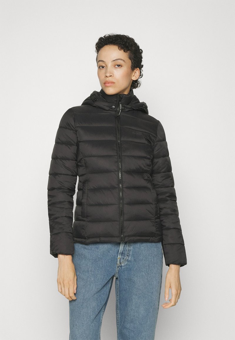 Chaqueta de plumas negra con cierre frontal, cuello alto y capucha ajustable. Presenta una textura acolchada y mangas largas, combinada con jeans de mezclilla azules.