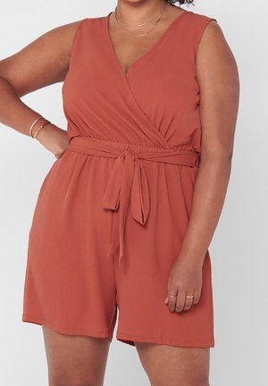 Femme portant une combinaison sans manches couleur rouille avec ceinture nouée à la taille et bijoux délicats en or au cou, au poignet et aux doigts.