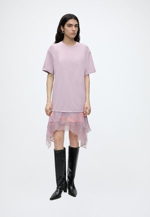 Kvinde med langt mørkt hår iført en lyse-lilla oversized T-shirt, gennemskinnet lyserød blondernederdel og sorte knæhøje støvler, står på en hvid baggrund.