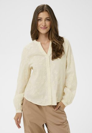 Kobieta o długich brązowych włosach, ubrana w kremową, haftowaną bluzkę i beżowe spodnie, uśmiechająca się, z jedną ręką w kieszeni, na białym tle.