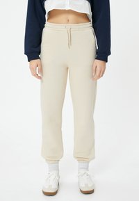 Beige Baumwoll-Sweatpants mit einem elastischen Bund und Bündchen. Verfügt über Seitentaschen und ein gerades Bein-Design. Kombiniert mit weißen Turnschuhen.