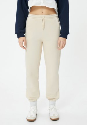 Beige katoenen sweatpants met een elastische tailleband en boorden. Voorzien van zijzakken en een recht model. G gedragen met witte sneakers.