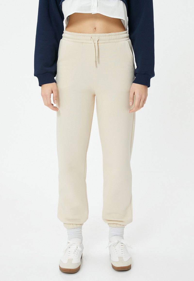 Beige Baumwoll-Sweatpants mit einem elastischen Bund und Bündchen. Verfügt über Seitentaschen und ein gerades Bein-Design. Kombiniert mit weißen Turnschuhen.